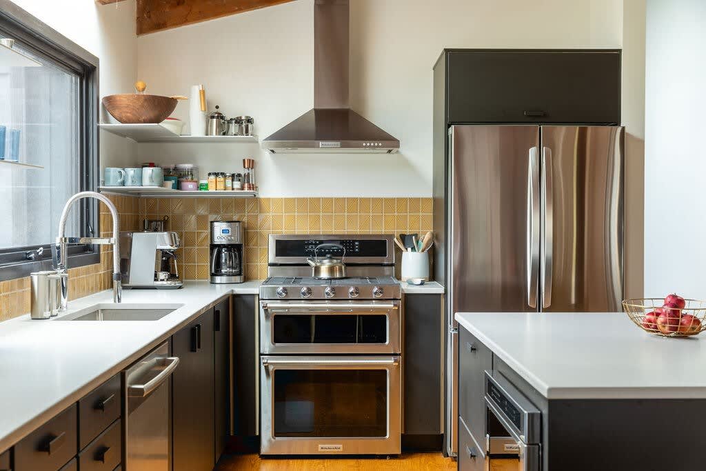 Modern kitchen with stainless steel appliances including a double oven range and refrigerator, white countertops, dark cabinetry, and a yellow tiled backsplash.