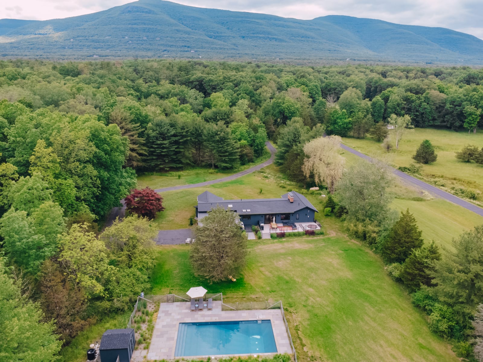 An aerial view showcases a dark-colored modern house nestled in a vast green landscape with dense forests and distant mountains. A rectangular swimming pool with a patio and a smaller outbuilding are visible in the foreground, connected by a winding driveway.