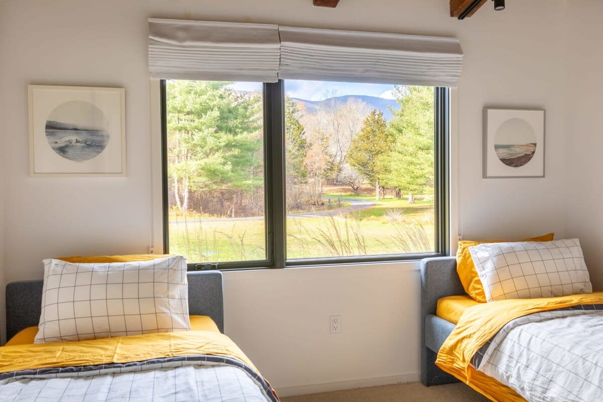 A bright bedroom featuring two twin beds with gray upholstered frames and yellow and white plaid bedding. A large window offers expansive views of a meadow and distant mountains.
