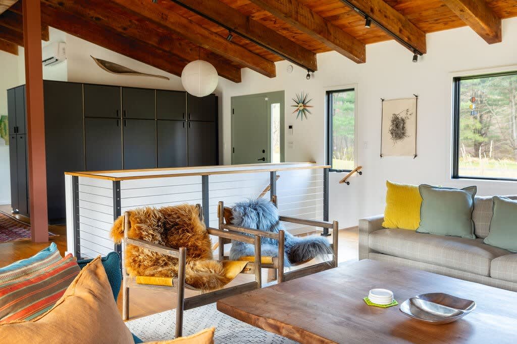 A spacious living area boasts an exposed wooden beam ceiling and track lighting, featuring a modern bar counter with dark cabinetry, a grey sofa with colorful pillows, and two unique chairs adorned with furry throws. Large windows provide ample natural light and outdoor views.