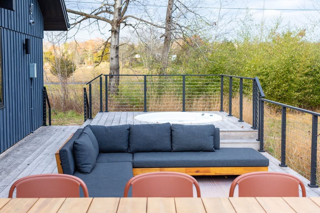 An expansive outdoor deck features a built-in hot tub and a modern L-shaped outdoor sofa with dark cushions, providing a comfortable seating area. The deck is enclosed by black metal railings and overlooks a natural, wooded landscape.