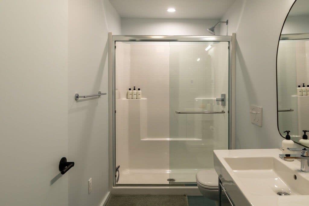 A modern bathroom features a glass-enclosed walk-in shower with white subway tiles, a sleek white vanity with a rectangular sink, and a large round mirror. The walls are painted a light blue, complemented by contemporary black fixtures.