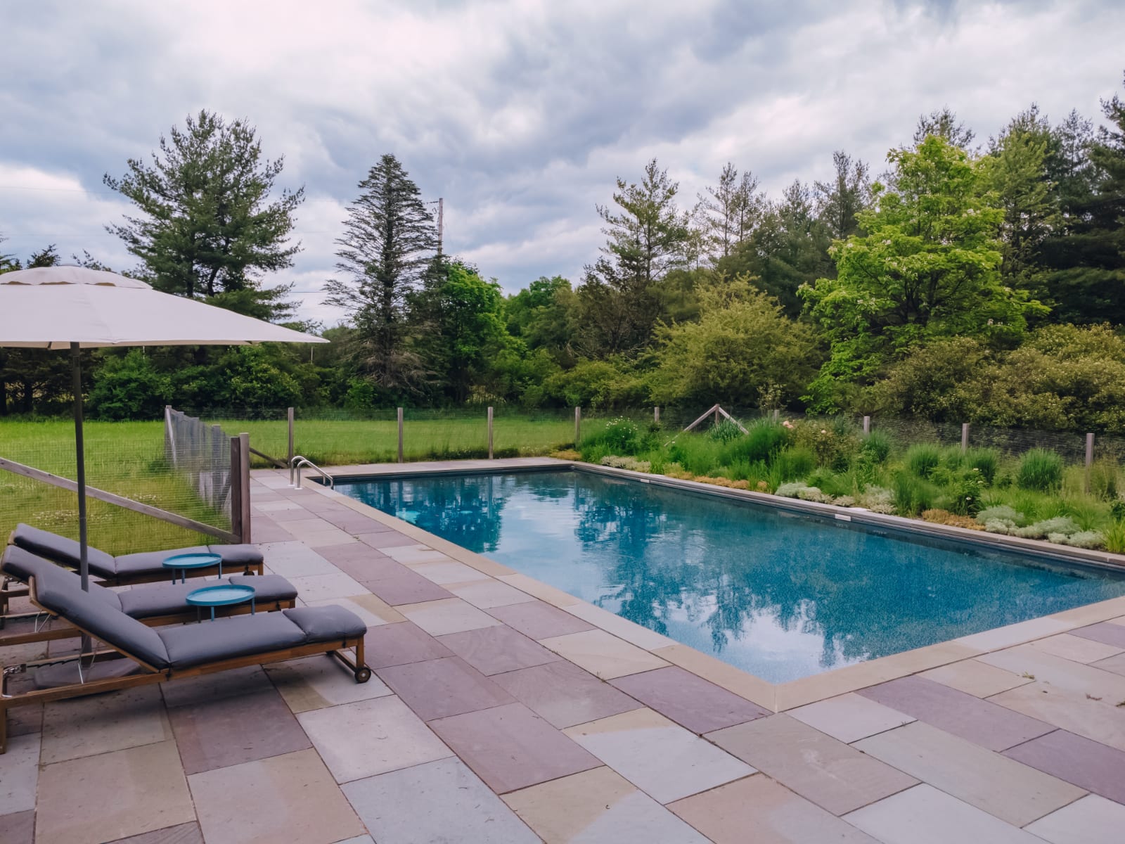 A clear blue rectangular swimming pool is surrounded by a multi-toned stone patio, with two grey lounge chairs and a white umbrella on one side. Lush green foliage and trees form the backdrop under a cloudy sky.