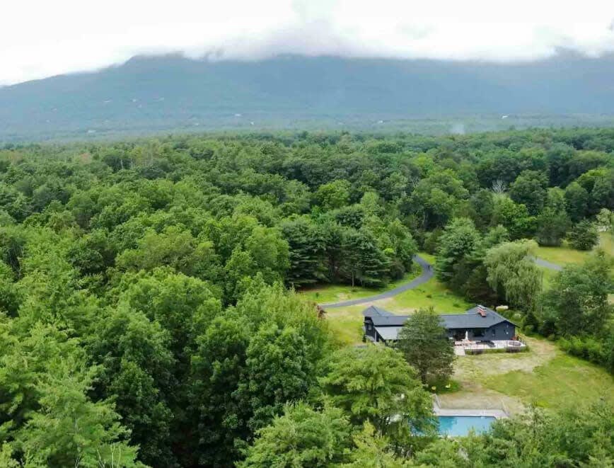 A high aerial view showcasing the dark gray house, swimming pool, and winding driveway amidst a vast expanse of dense green forest. Distant mountains are visible under a cloudy sky, emphasizing the secluded, rural setting.