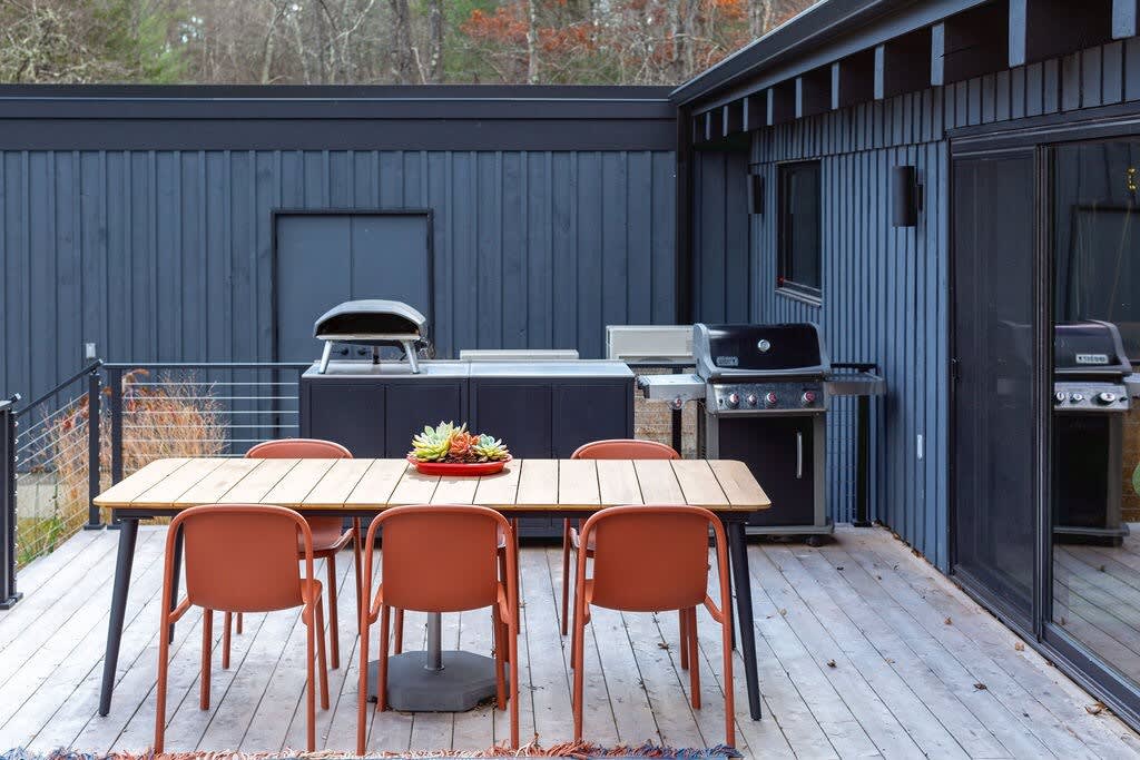 An outdoor dining area on a wooden deck features a long wooden table with four orange chairs, set against the dark blue exterior of the house. A built-in outdoor kitchen with a grill and pizza oven is visible in the background, surrounded by trees.