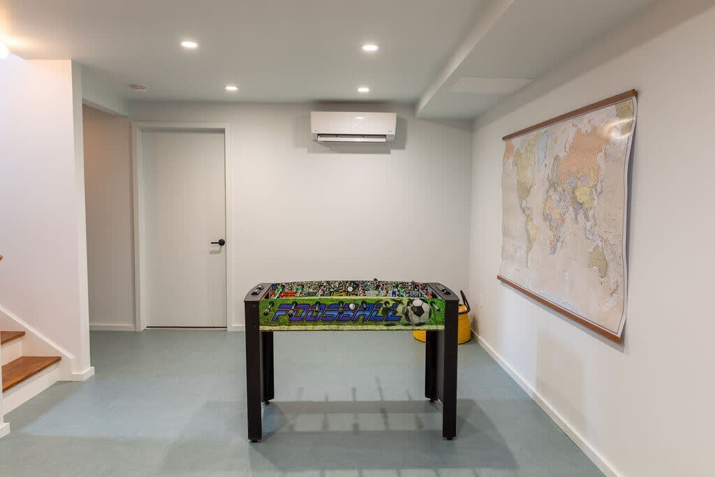 A minimalist game room features a foosball table as its central element, set on a light blue floor with clean white walls and recessed lighting. A large world map hangs on one wall, and an air conditioning unit is mounted above a white door.