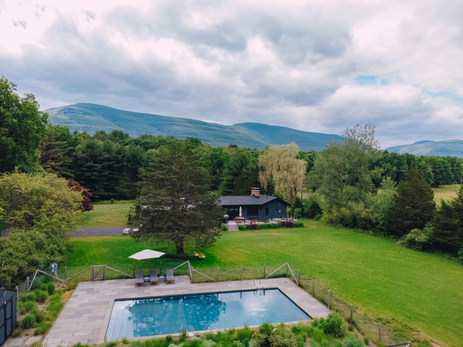 An aerial view of the dark gray modern house nestled in a lush green landscape with a rectangular swimming pool and lounge chairs. Rolling mountains and a cloudy sky form a picturesque backdrop.