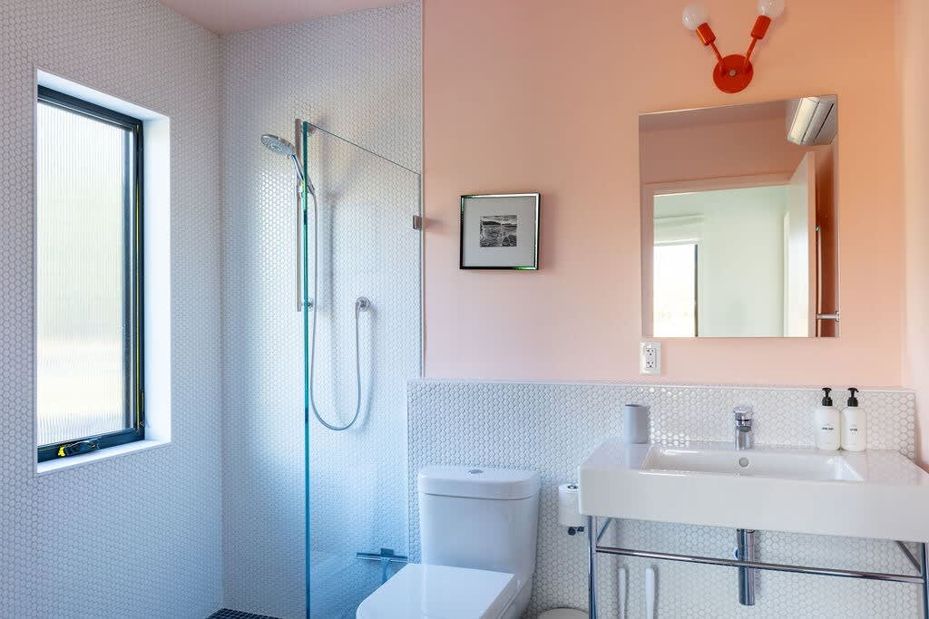 A modern bathroom with white honeycomb tile walls and a light pink painted upper wall. It features a white toilet, a vanity with a sink, a mirror, and a glass-enclosed shower with a rainfall showerhead.