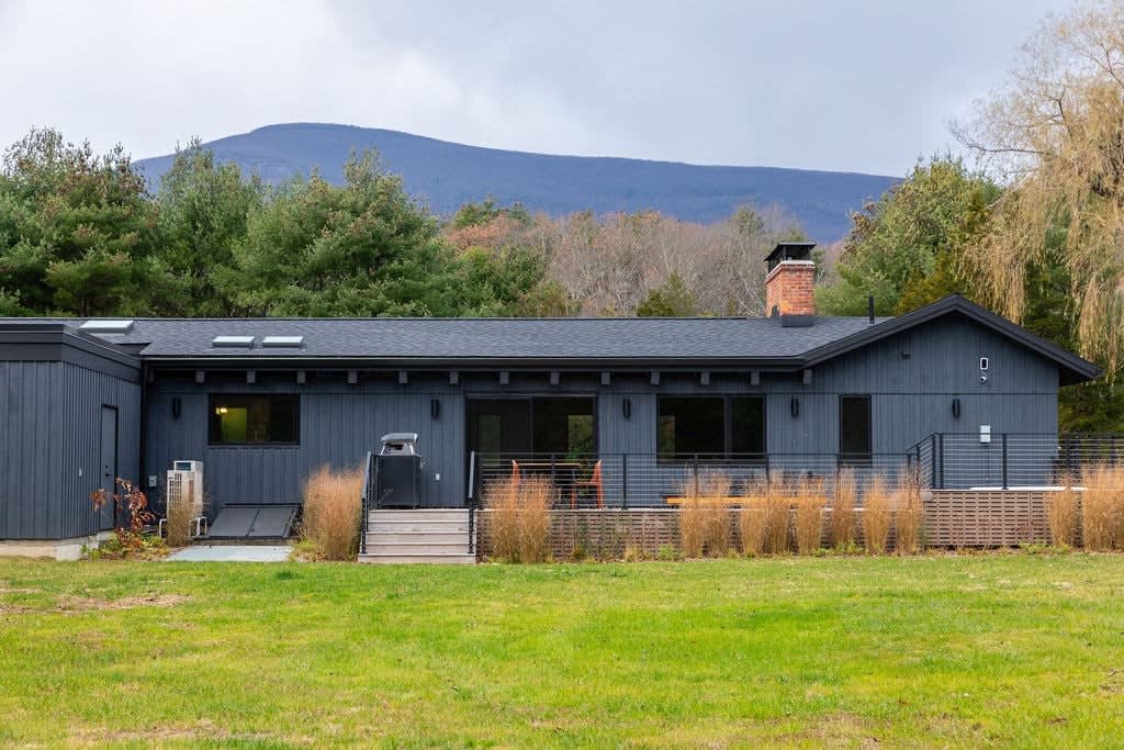 The back exterior of a modern dark grey house with a black roof, featuring multiple large windows and a spacious deck. A brick chimney adds a classic touch, and the house is set against a backdrop of trees and a distant mountain range under a cloudy sky.