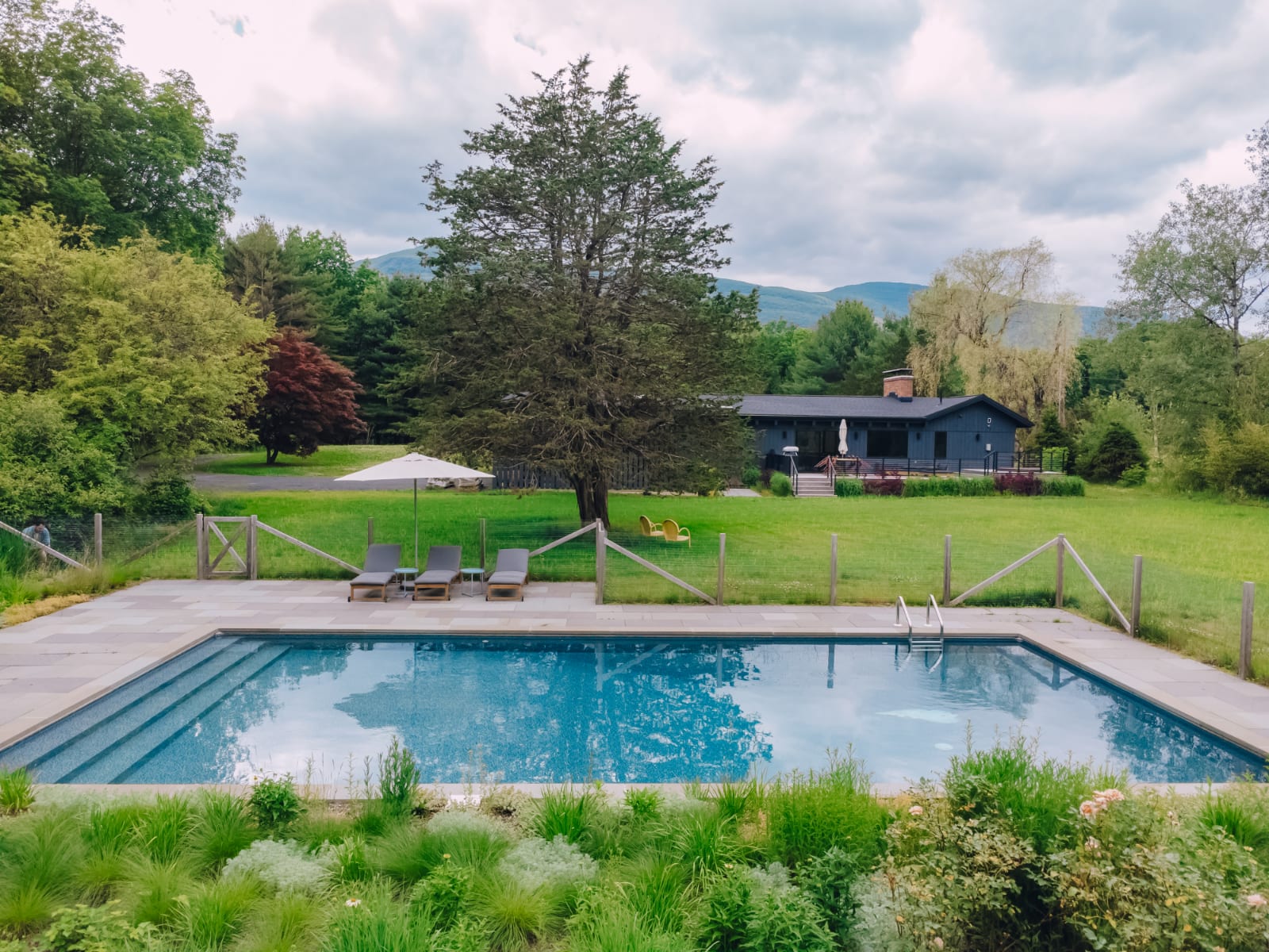 An aerial view of the property showcases a large blue swimming pool surrounded by a stone patio and a sprawling green lawn. The dark grey house is visible in the background, nestled among mature trees with mountains in the distance under a partly cloudy sky.
