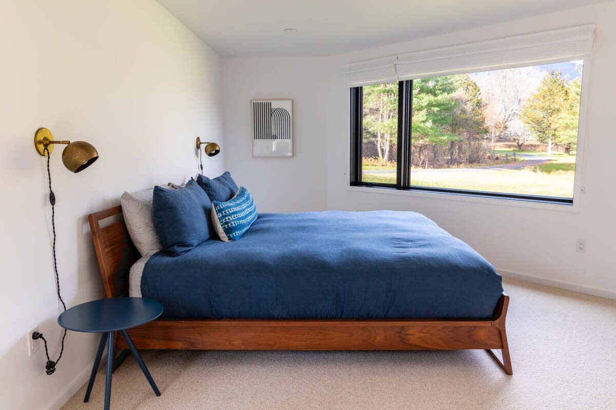 A serene bedroom features a wooden bed frame with a deep blue duvet and matching pillows, flanked by brass wall sconces and a small blue side table. A large window offers expansive views of trees and distant scenery, with a piece of abstract art on the wall.