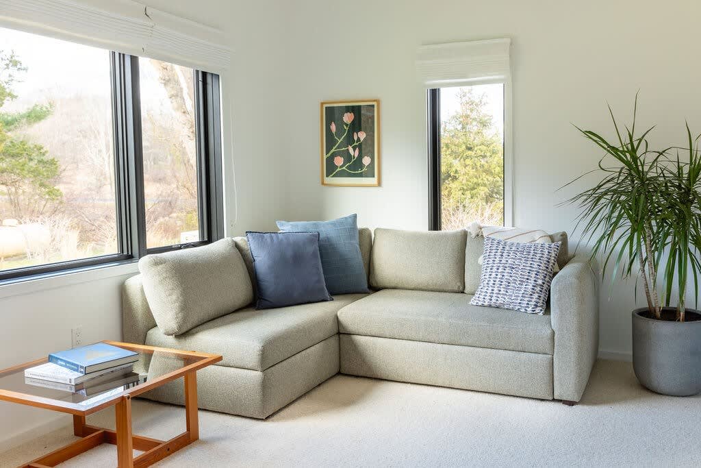 A cozy living room features a light gray L-shaped sectional sofa with blue throw pillows, a glass-top coffee table with books, and large windows offering views of lush greenery. A potted plant sits in the corner, and a framed botanical print hangs on the wall.