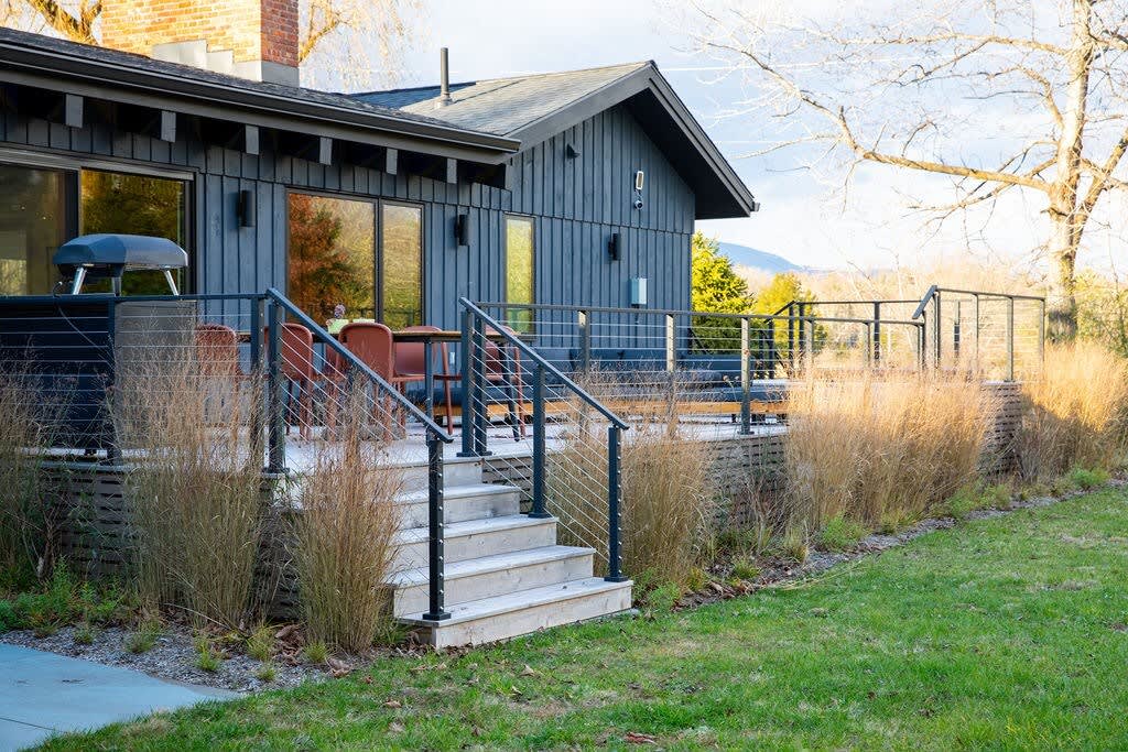 A spacious wooden deck extends from the dark gray house, furnished with an outdoor dining set and a BBQ grill. Modern black cable railings enclose the deck, which is bordered by tall ornamental grasses and overlooks a grassy yard.