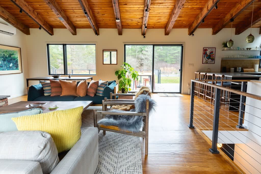 Bright, open-plan living room with exposed wooden beams, hardwood floors, multiple seating areas including a grey sofa and chairs with faux fur throws, and a sliding glass door leading to the outdoors.