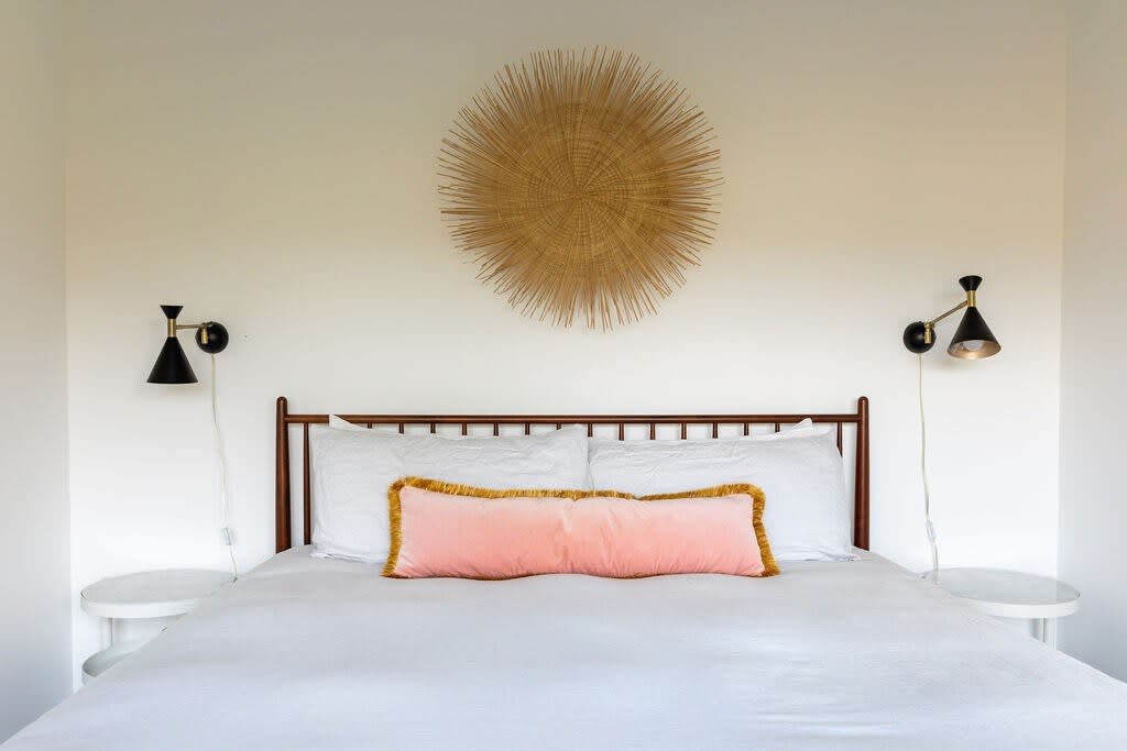 Serene bedroom featuring a bed with crisp white linens, a wooden headboard, two black wall sconces, and a large circular woven wall hanging above the bed.