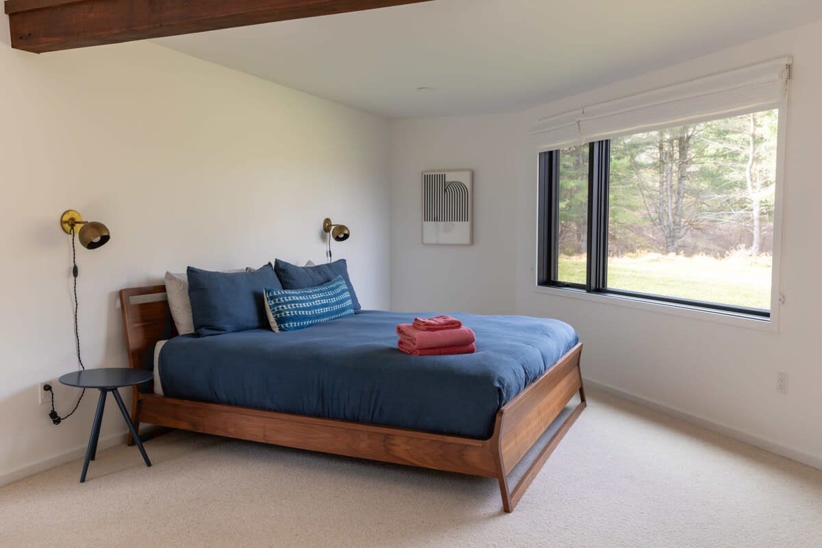 A spacious bedroom with a wooden bed frame, dark blue bedding, and two small side tables with wall-mounted brass sconces. A large window offers views of the outdoors, and a minimalist framed print hangs above the bed.