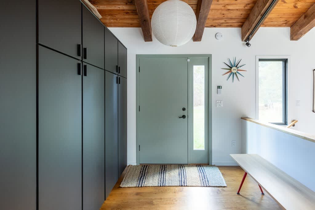 A welcoming entryway with a light green front door, dark gray floor-to-ceiling storage cabinets, and a wooden bench. The ceiling features exposed wooden beams and a large white spherical pendant light.
