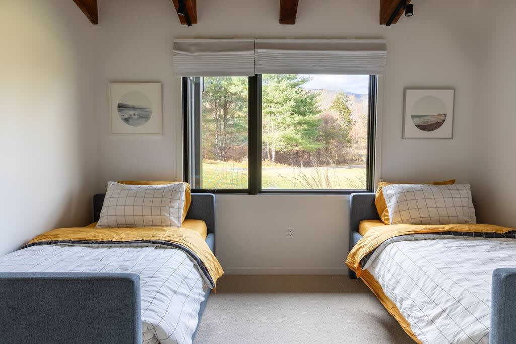 A bright bedroom features two twin beds with yellow and white checkered bedding, positioned beneath a large window offering scenic views of lush greenery and distant mountains. The room has light walls, a neutral carpet, and framed artwork.