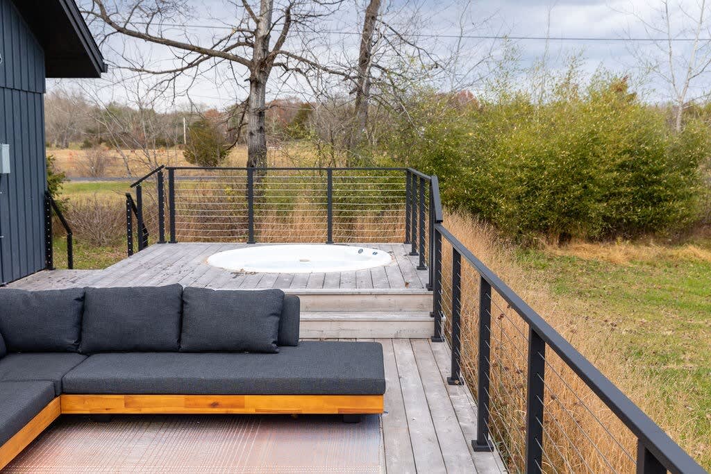 A wooden deck features a built-in white hot tub and a dark gray outdoor sectional sofa with wooden base. Modern black cable railings enclose the deck, which overlooks a natural, grassy landscape with trees.