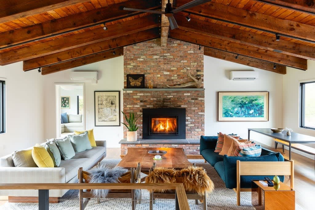 A cozy living room featuring a prominent brick fireplace with a black insert, flanked by two sofas with colorful pillows. The room boasts a vaulted wooden beam ceiling with a ceiling fan and track lighting, and a large wooden coffee table anchors the space.