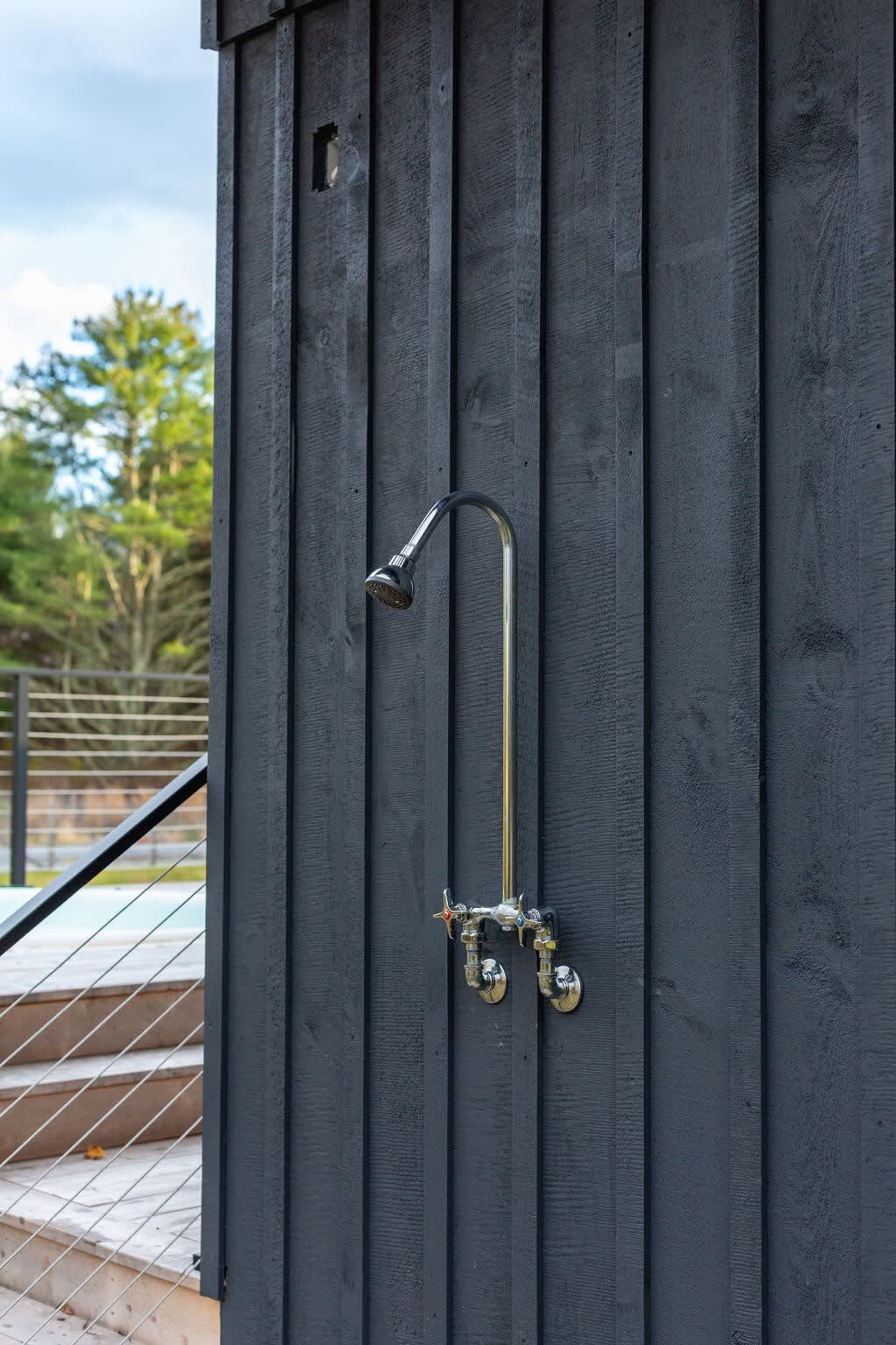 A close-up of the dark gray vertical board-and-batten exterior wall of the house, featuring a modern chrome outdoor shower fixture with exposed pipes and handles. A deck with cable railings is visible in the background.