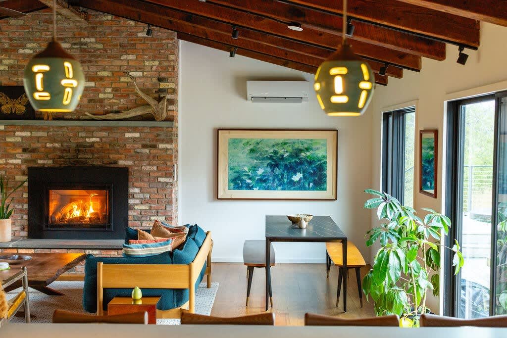 View of an open-concept living space showcasing a brick fireplace, exposed wooden beams, a blue sofa, and a black dining table with benches, illuminated by decorative pendant lights.