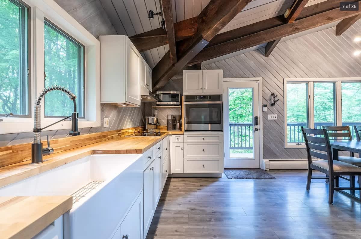 Bright kitchen featuring white shaker cabinets, butcher block countertops, a large white farmhouse sink with a gooseneck faucet, and a door leading to an outdoor deck.