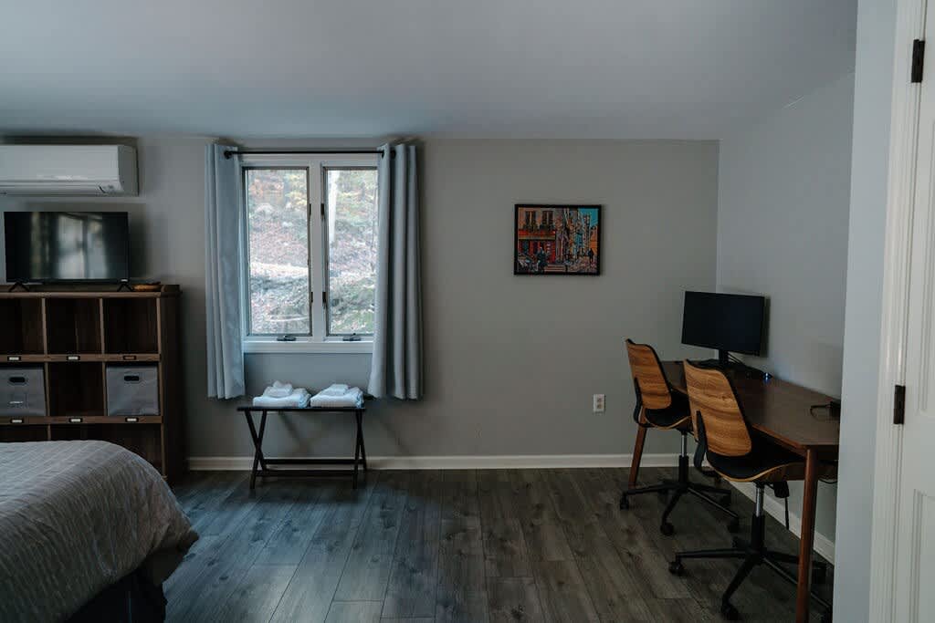 A bright room features a bed partially visible on the left, a TV on a wooden storage unit, and a window with light gray curtains. A dedicated workspace with a desk, two chairs, and a computer monitor is set against a light gray wall.