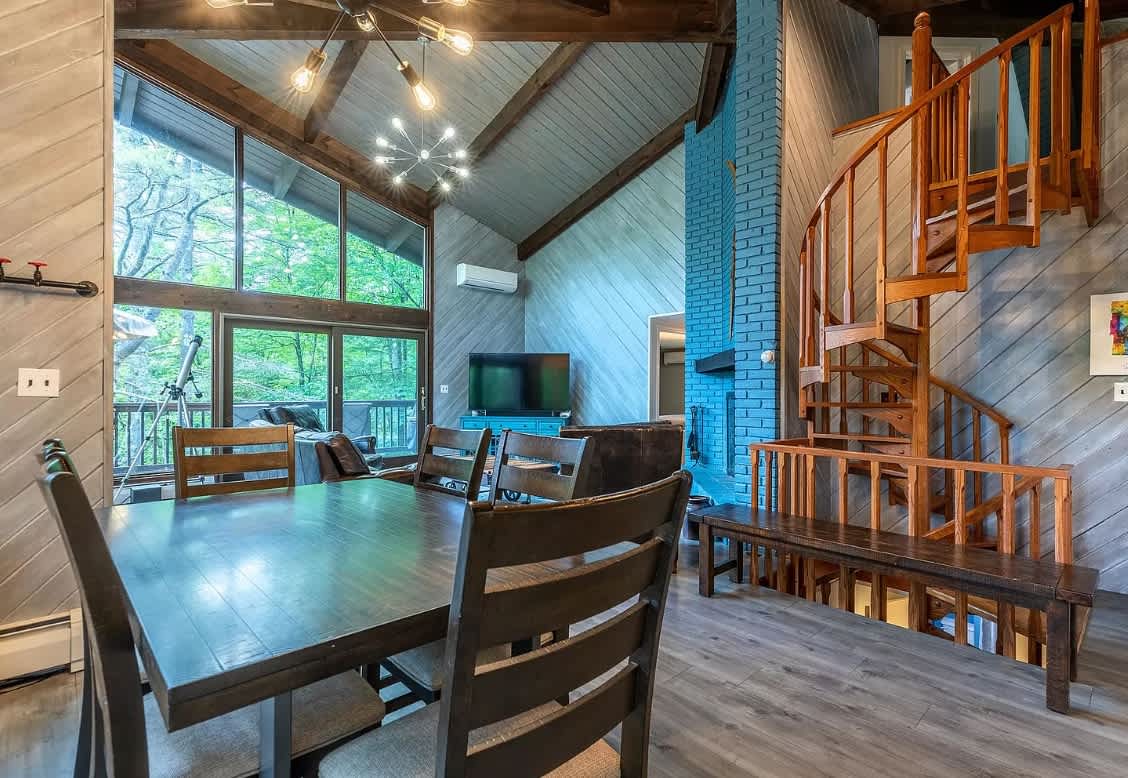 Spacious dining area with a dark wooden table and chairs, featuring a striking wooden spiral staircase and an open view into the living room with its large windows and vaulted ceiling.