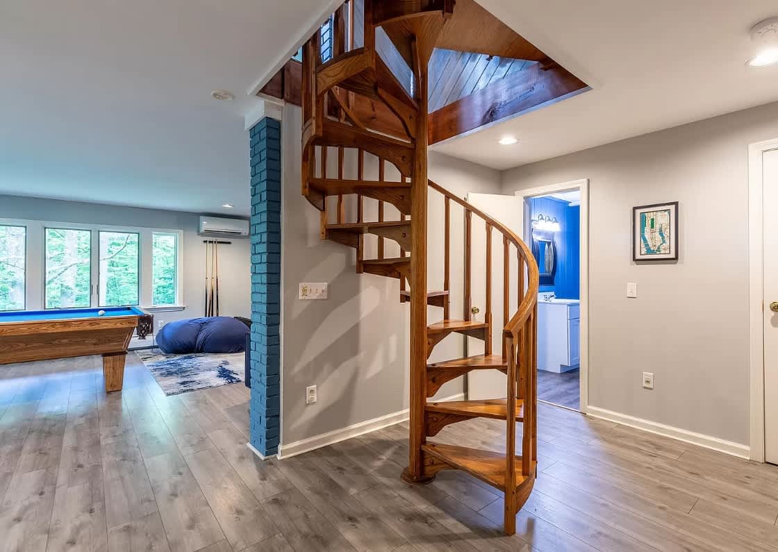 Open game room with a prominent wooden spiral staircase, light gray walls, and wood-look flooring. A blue brick column adds a pop of color, and a doorway leads to a bathroom.
