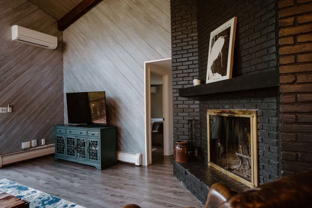 A rustic living room showcases a black brick fireplace with a wooden mantel, a TV on a teal console, and a sloped ceiling with light-colored wooden planks. A white air conditioning unit is mounted on the wall, and a large artwork hangs above the fireplace.