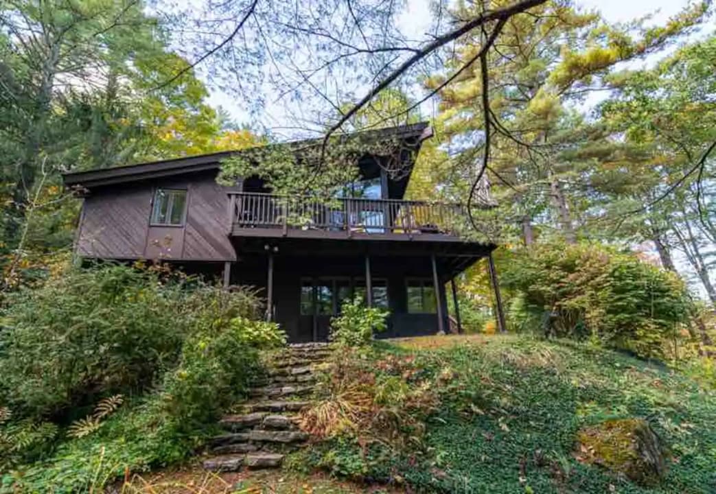 A dark brown A-frame style house nestled on a lush green hillside, surrounded by trees with some autumn foliage, featuring a large deck with a railing and stone steps leading up to the entrance.