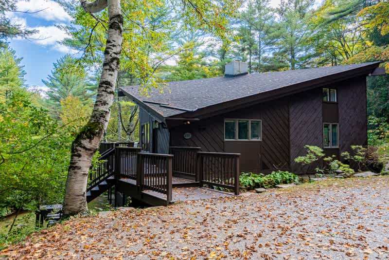 The front exterior of a dark brown modern cabin with a sloped roof, featuring a wooden deck leading to the entrance and a gravel driveway surrounded by trees with autumn leaves.