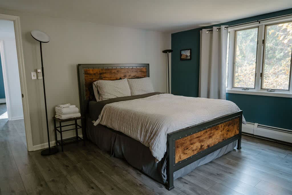 A cozy bedroom features a bed with a dark metal and wood frame, light bedding, and a dark accent wall. A tall floor lamp stands next to a small black metal nightstand with folded towels, and a large window offers natural light.