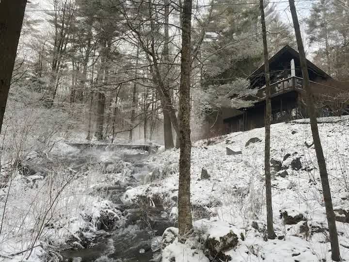 A dark brown house nestled in a snow-covered forest during winter, with a misty stream flowing in the foreground and bare trees covered in frost, showcasing the property's seasonal charm.
