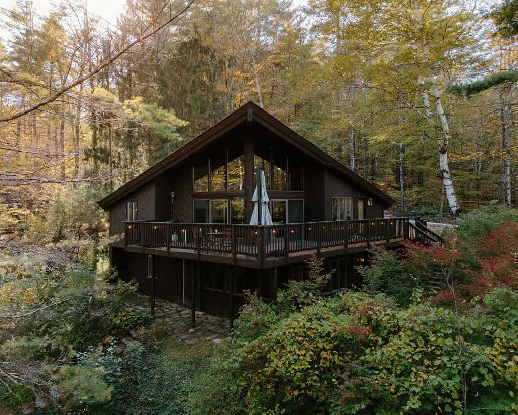 A striking dark brown A-frame house with a prominent deck and expansive floor-to-ceiling windows, nestled amidst a vibrant autumn forest.