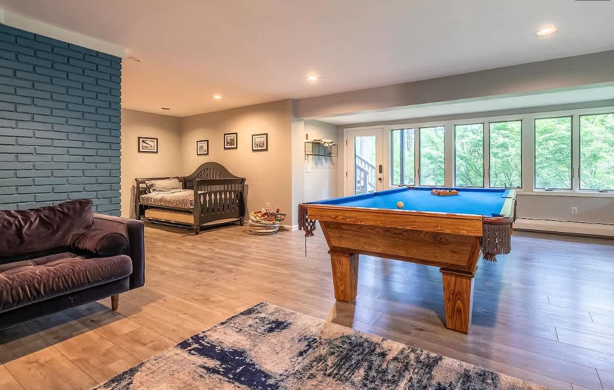 Wide view of the game room with a dark velvet sofa, a daybed, and a pool table. A blue brick wall adds texture, and large windows provide ample natural light.