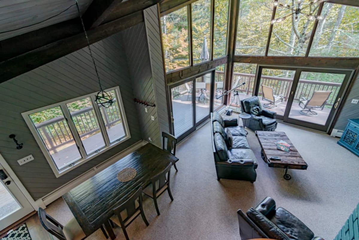 Aerial view of the open-concept living and dining area, highlighting the vaulted ceiling, expansive windows, and the seamless flow between the seating area and the dark wooden dining table.