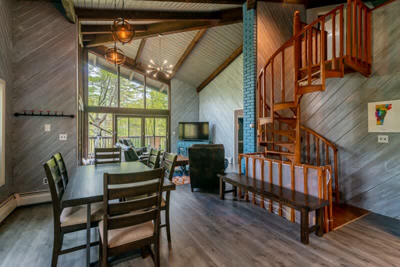 Open-concept dining area with a dark wooden table and chairs, adjacent to the living room with large windows, and featuring a prominent wooden spiral staircase leading to an upper level.