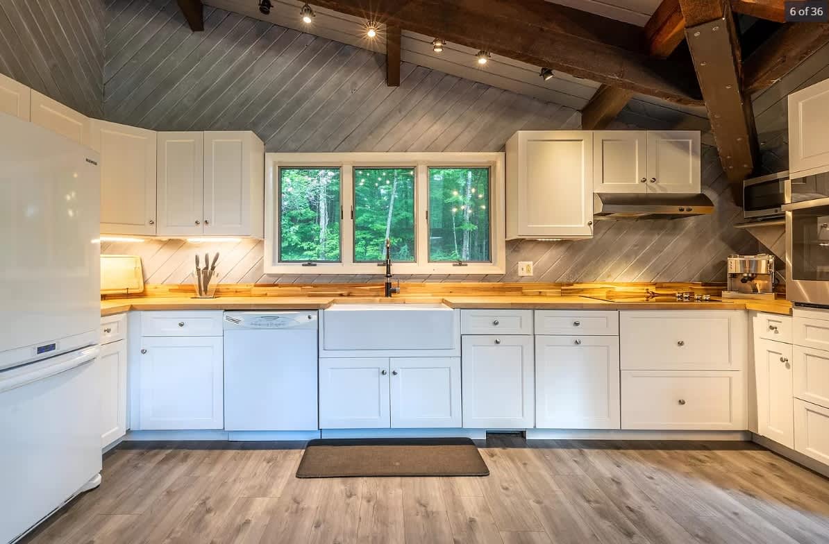 Modern kitchen with white shaker cabinets, light wood countertops, a large farmhouse sink, and stainless steel appliances. The ceiling features exposed wooden beams and white slatted wood.