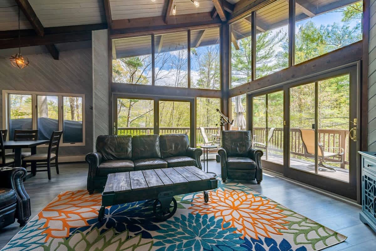 Expansive living room with soaring windows showcasing the surrounding greenery, furnished with a dark leather sofa, a unique wooden coffee table, and a vibrant floral area rug.