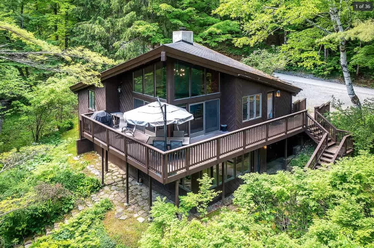 An aerial view of a dark brown modern cabin with a large wraparound deck, nestled among dense green trees, featuring an outdoor dining area with an umbrella.