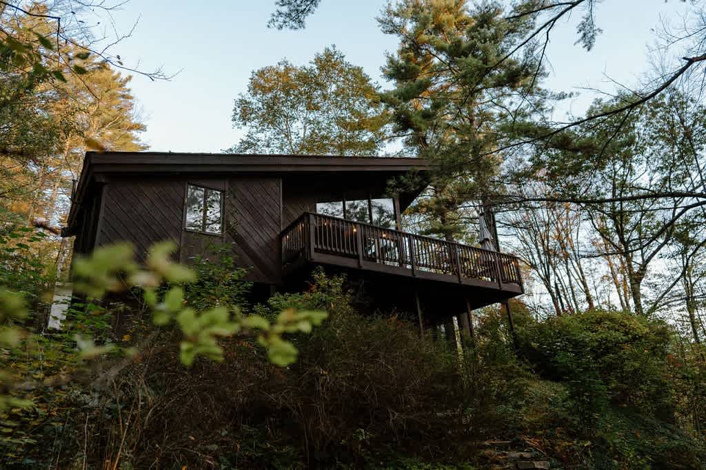 A dark brown modern cabin with a large deck, partially obscured by lush green trees and foliage, viewed from a lower angle on a hillside, highlighting its secluded setting.