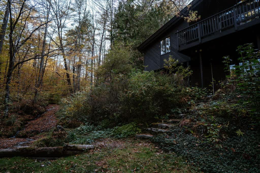 The dark-sided house is nestled into a lush, wooded hillside with a small stream flowing through the autumn-colored trees. Stone steps lead up to the house, which features a dark balcony overlooking the natural landscape.