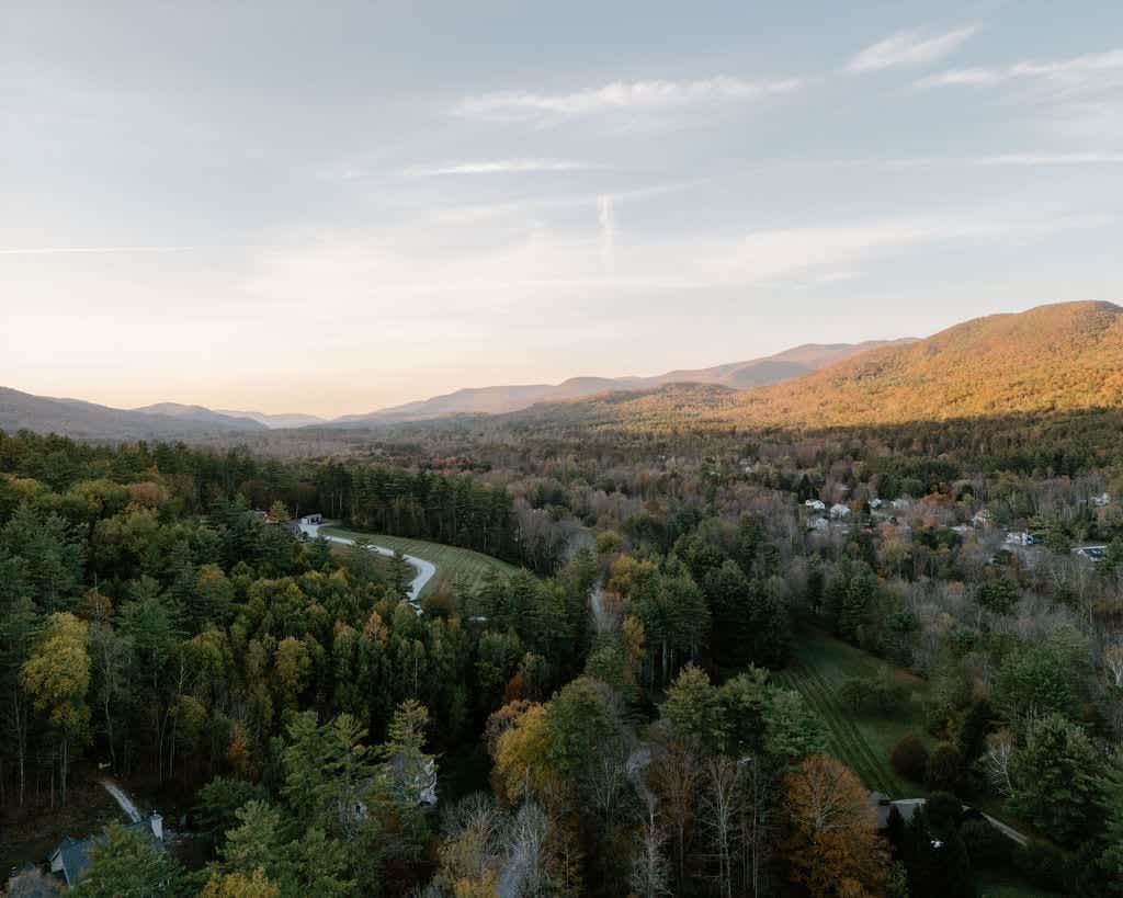 A breathtaking aerial view of a vast valley filled with dense forests displaying autumn colors, with rolling mountains in the distance under a soft, cloudy sky.