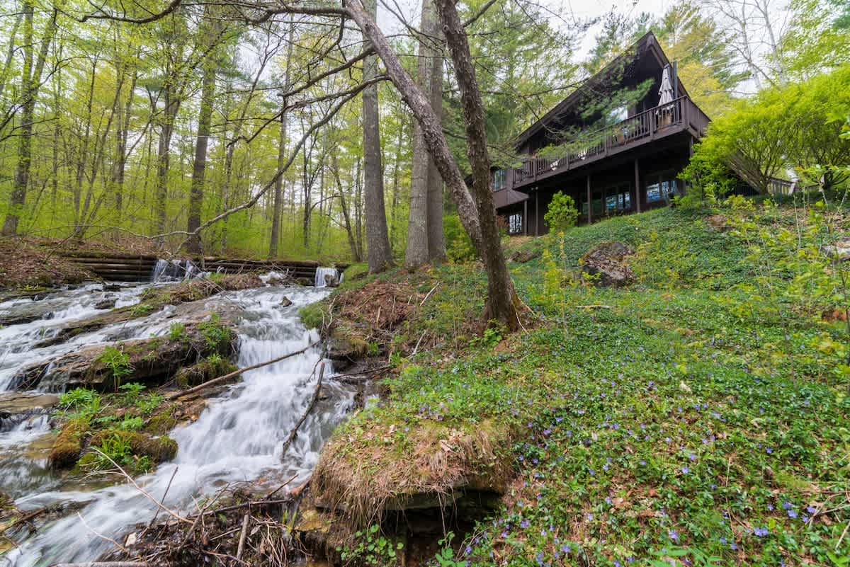 The dark A-frame house is perched on a lush green hillside, overlooking a picturesque cascading stream with small waterfalls, surrounded by dense forest.