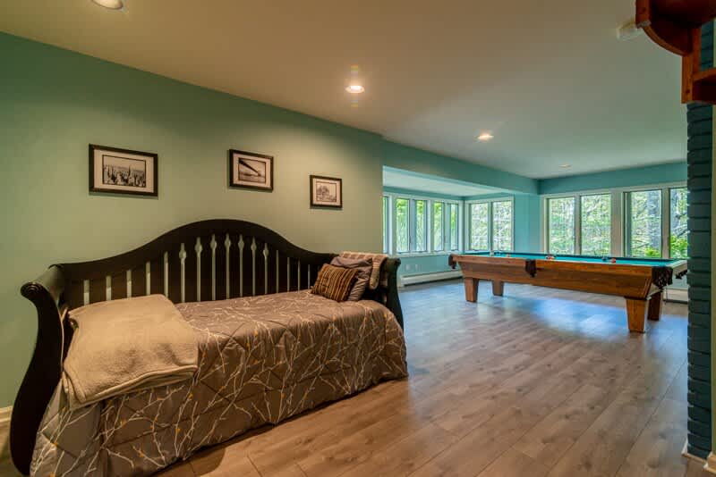 Spacious game room featuring a daybed with a dark wooden frame and a pool table with a blue felt top. Large windows offer views of the outdoors, and the walls are painted light blue/green.