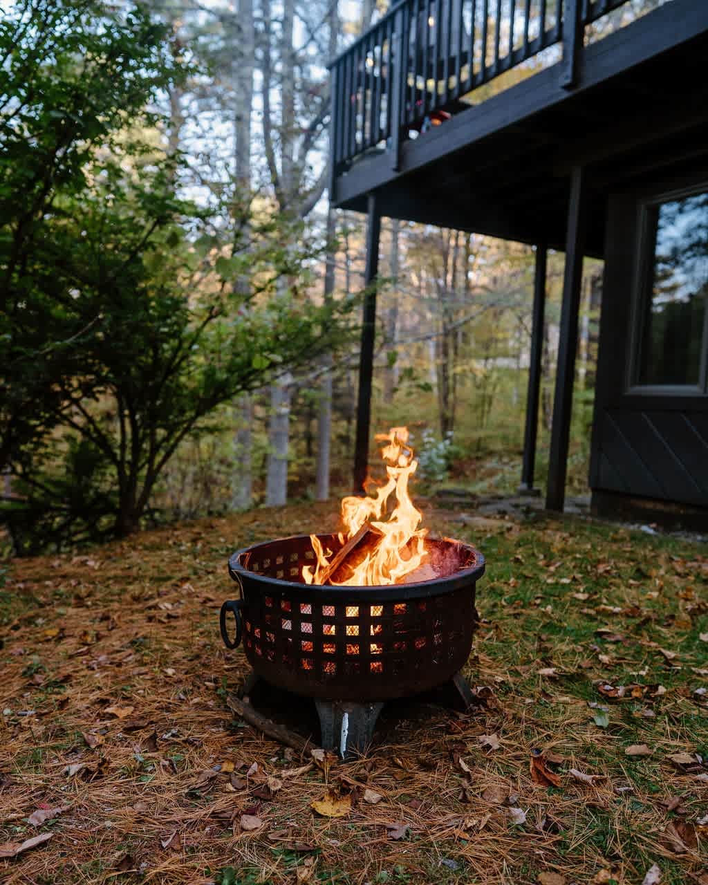 A metal fire pit with a decorative cut-out pattern burns brightly with logs, surrounded by fallen leaves and pine needles. In the background, a dark house with a deck is visible amidst autumn trees.