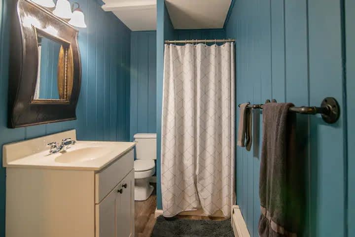 A bathroom featuring teal blue vertical paneling on the walls, a white vanity with a light countertop, a rectangular mirror, and a white shower curtain with a subtle geometric pattern.
