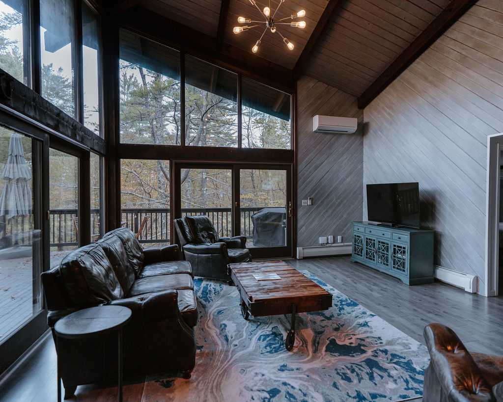 Bright living room with a vaulted wood-paneled ceiling, large floor-to-ceiling windows offering forest views, a dark leather sofa, and a rustic wooden coffee table on a blue and white abstract rug.